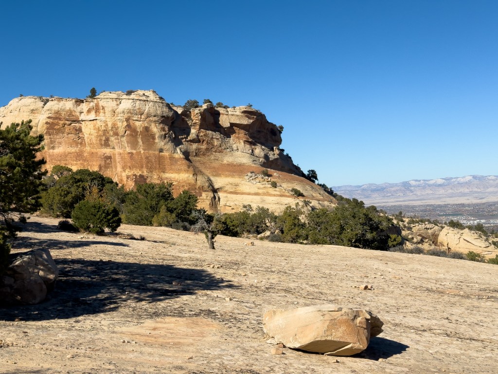 Sunlit sandstone cliff with layered red and tan rock formations in Colorado National Monument, surrounded by scattered green shrubs and trees on a rocky plateau under a clear blue sky, with distant mountains visible on the horizon.