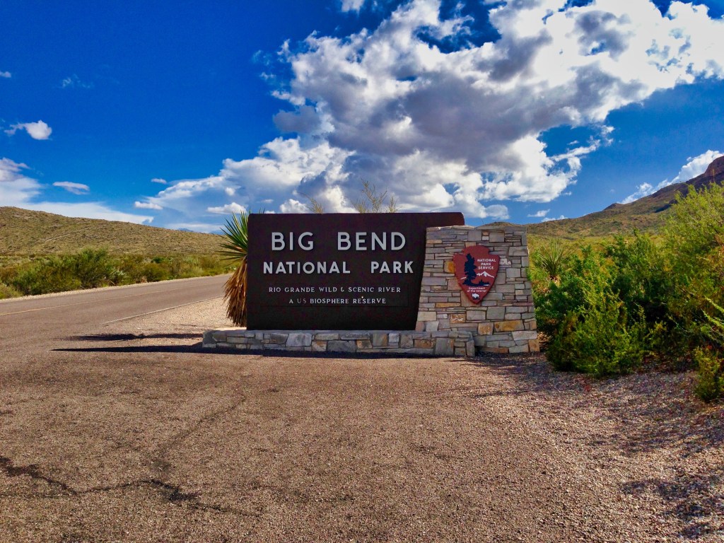 Entrance sign for Big Bend National Park along a paved road, featuring a large brown panel with white lettering and a stone base. The sign includes the National Park Service emblem and text noting “Rio Grande Wild & Scenic River” and “A U.S. Biosphere Reserve.” Surrounding the sign are desert plants, rugged hills, and a partly cloudy blue sky.